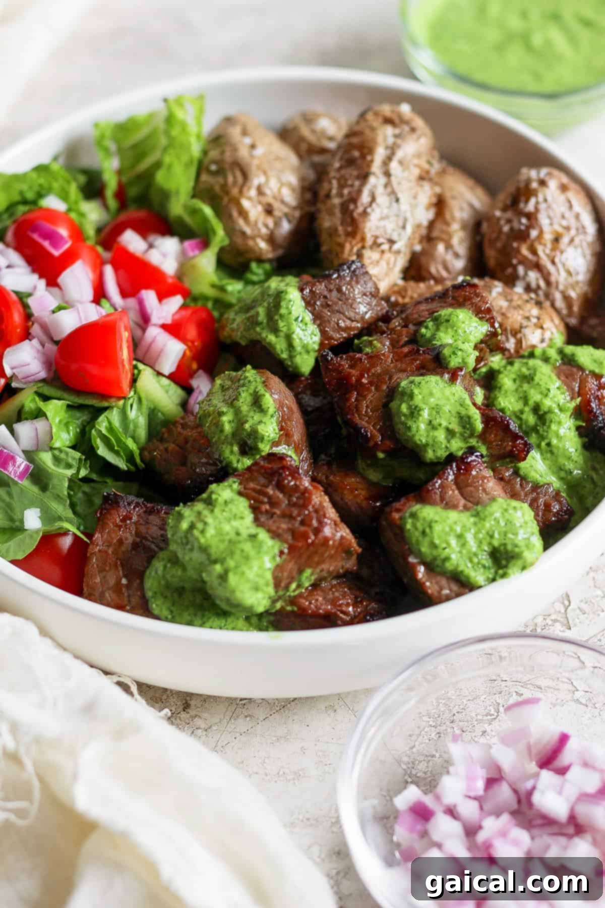A complete and inviting bowl featuring grilled steak, a generous topping of green chimichurri sauce, roasted potatoes, and a fresh salad of lettuce, cherry tomatoes, and red onions. A small dish of minced red onions accompanies the bowl.