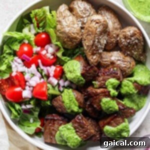 A beautifully arranged bowl containing golden roasted potatoes, a fresh salad with crisp lettuce, juicy tomatoes, and red onions, and tender air fryer steak bites generously topped with a vibrant green sauce.