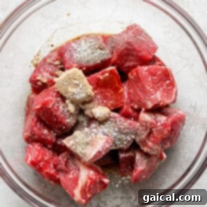 A glass bowl filled with raw beef chunks seasoned with spices on top, placed on a light-colored textured surface.