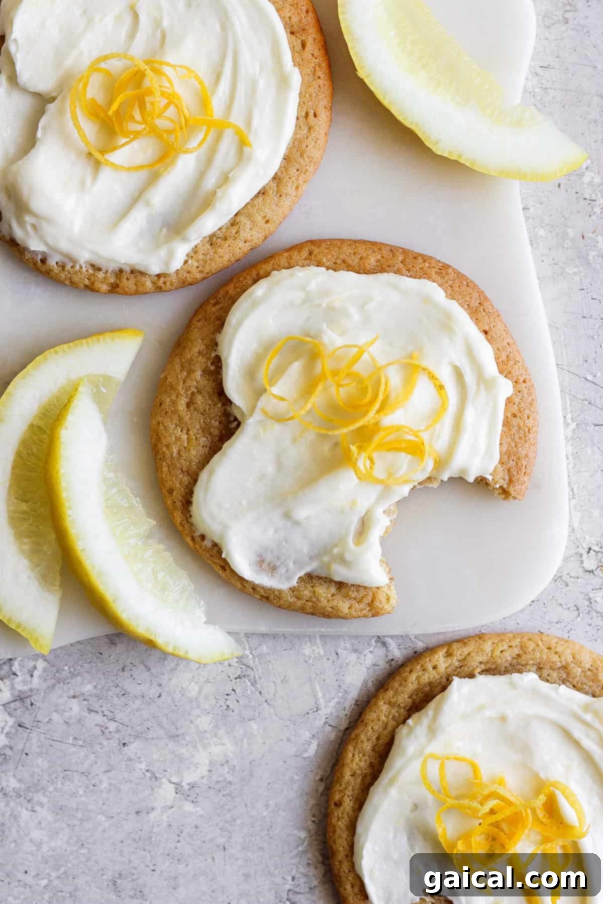 Three lemon cookies with cream cheese frosting, topped with fresh zest, sit invitingly on a cutting board. One is partially eaten, revealing its soft interior. Beside them, lemon wedges are artfully arranged on the textured surface, enhancing the citrusy allure of the scene.