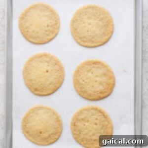 Six round lemon cookies with cream cheese frosting rest on a parchment-lined baking sheet.