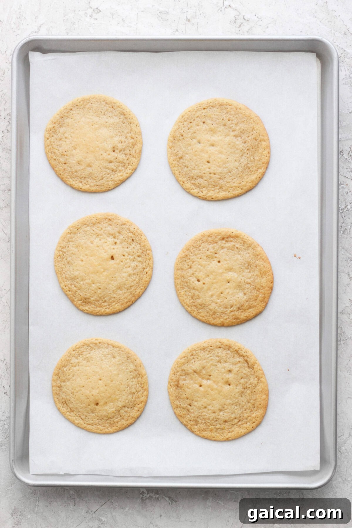 Six round lemon cookies with cream cheese frosting rest on a parchment-lined baking sheet.