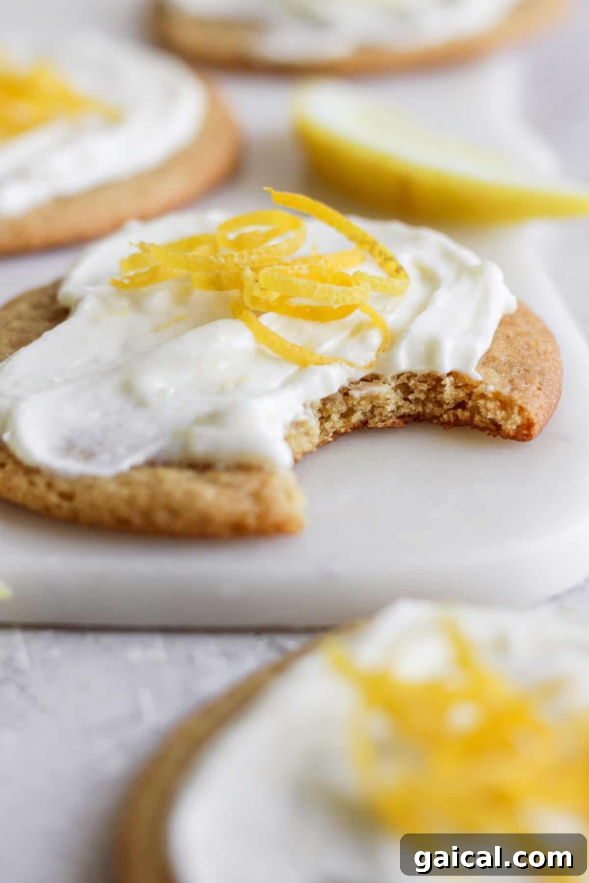 Close-up of a lemon cookie with cream cheese frosting and lemon zest on top. A bite is taken out of the cookie. A lemon wedge is visible in the background.