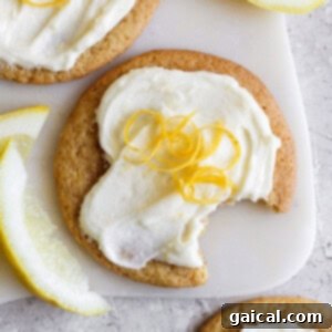 A lemon cookie with cream cheese frosting, topped with lemon zest, has a bite taken out of it. Slices of lemon are nearby on a light surface.