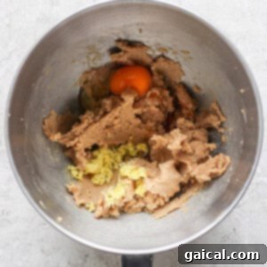 A mixing bowl brimming with cookie dough, a cracked egg, grated ginger, and hints of lemon zest prepares the stage for delightful lemon cookies with cream cheese frosting on a light textured surface.