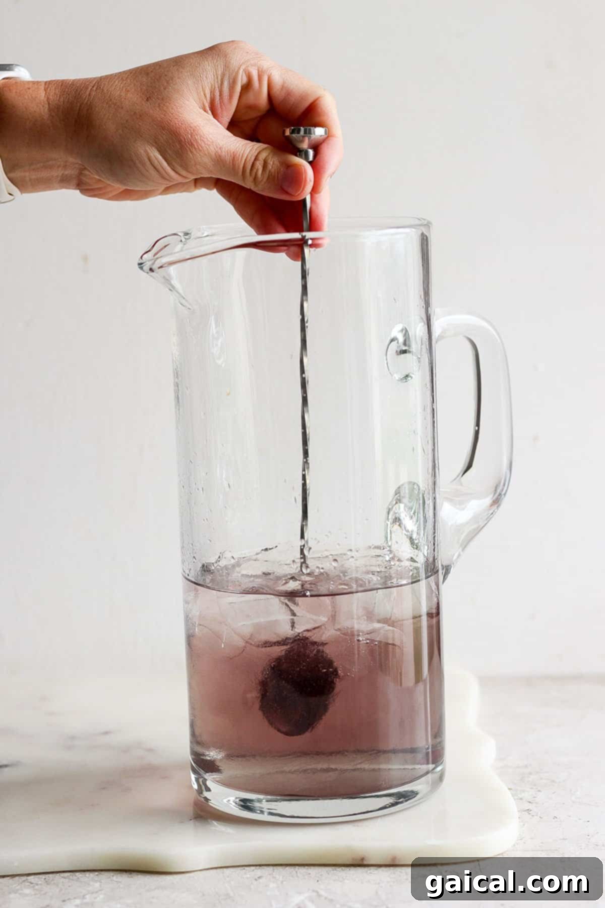 A close-up of a hand stirring a purple liquid in a clear pitcher, illustrating the infusion process.