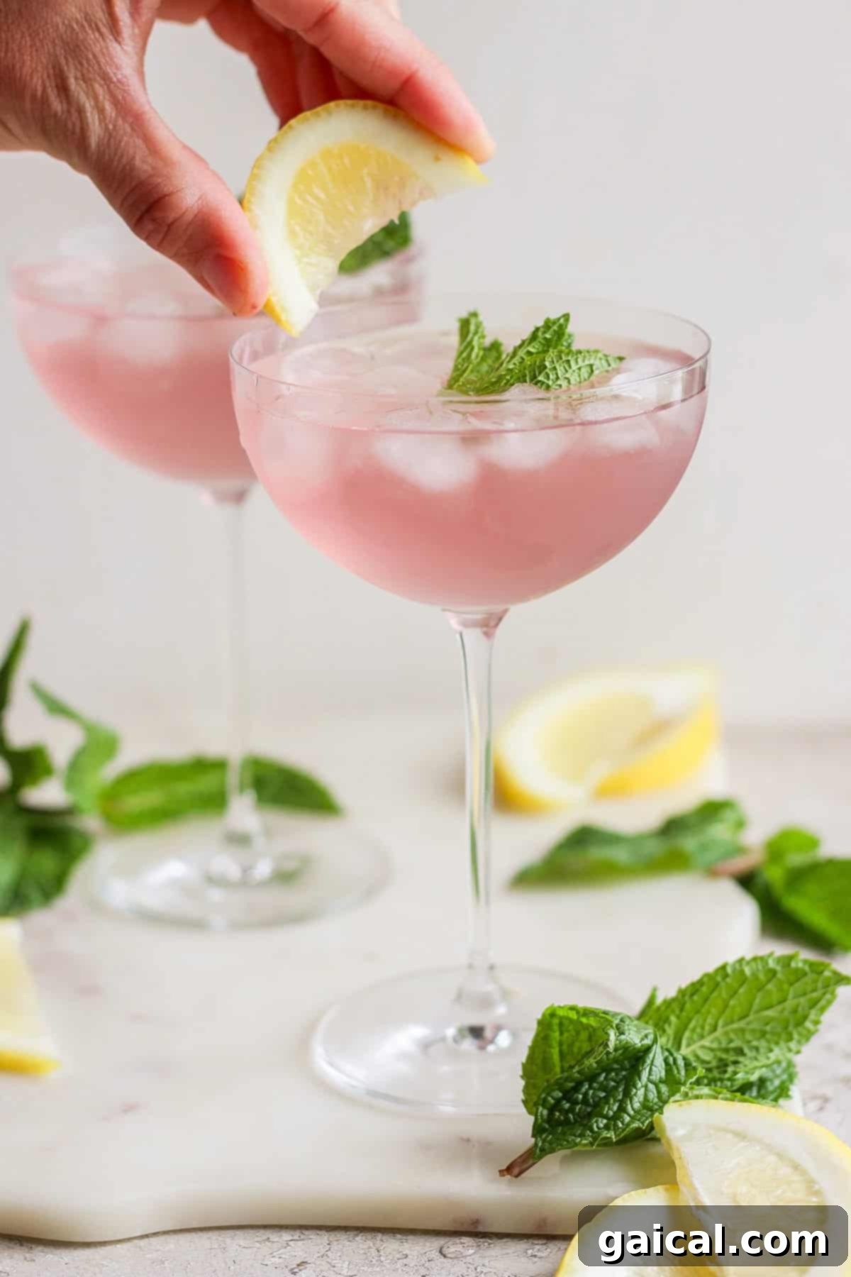 A hand placing a lemon wedge on the rim of a glass filled with pink lemonade garnished with mint leaves, with another similar glass in the background, showcasing the transformed color.