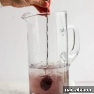 A hand stirring a purple liquid with ice in a clear glass pitcher using a metal spoon.