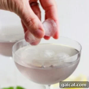 A hand pouring a cocktail into a glass with ice, with other glasses blurred in the background.
