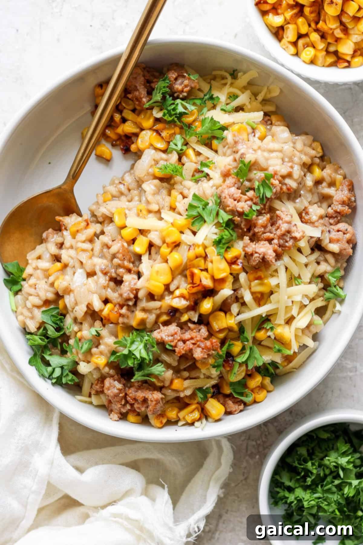 A beautifully presented bowl of creamy sweet corn risotto, generously topped with cooked ground Italian sausage, golden corn kernels, and shredded cheese, garnished with vibrant fresh parsley. A golden spoon rests invitingly on the side of the bowl. A smaller bowl of fresh corn is blurred in the background, hinting at the dish's star ingredient.