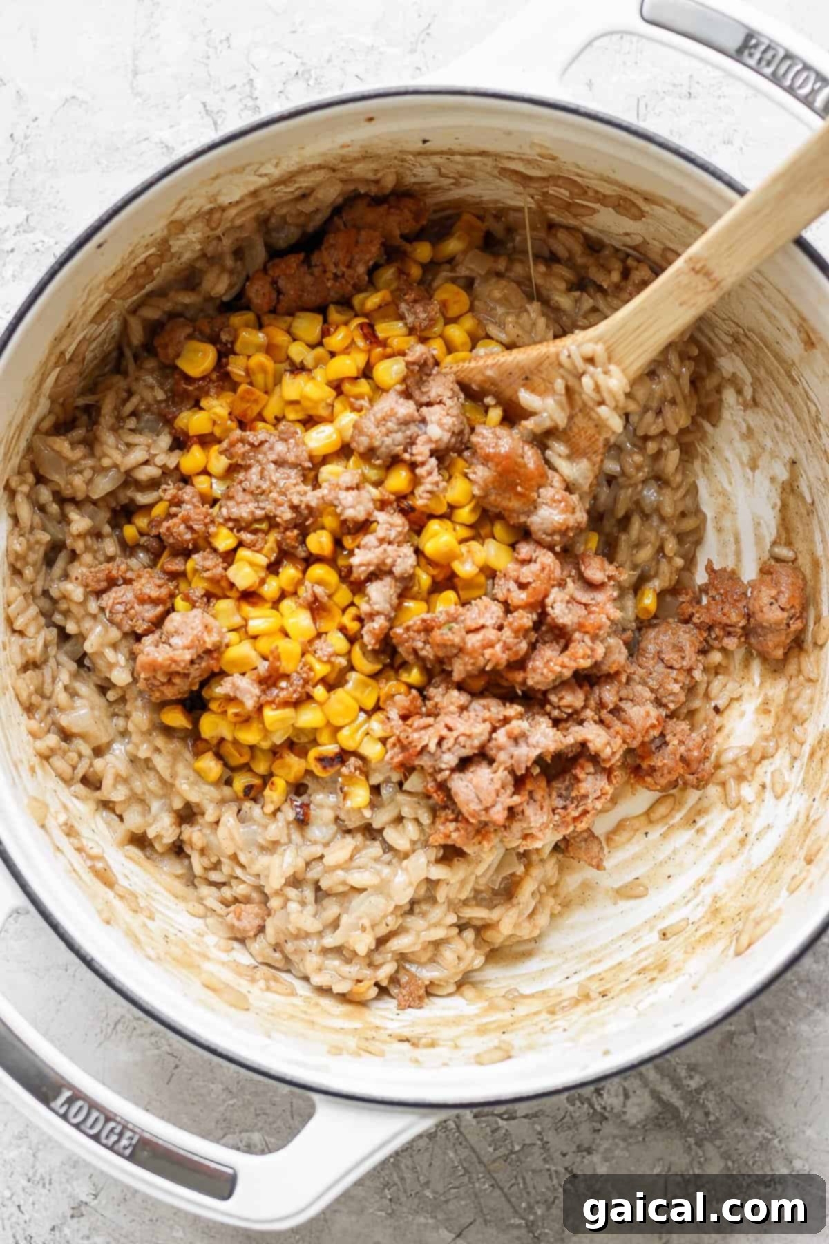 A large white Dutch oven on a textured grey surface, containing creamy risotto with visible kernels of corn and pieces of ground Italian sausage, being stirred gently with a wooden spoon.