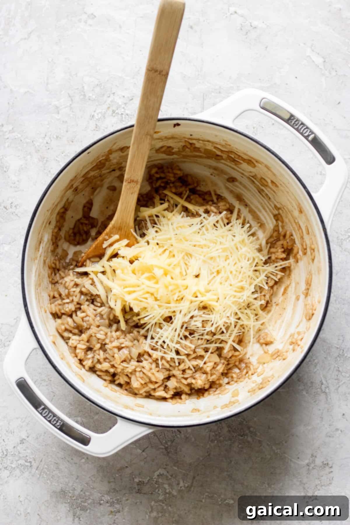 A rustic white pot on a light grey textured surface holds creamy risotto. Freshly grated cheese, a mix of Gouda and Parmesan, is piled atop the risotto, ready to be stirred in with a wooden spoon resting nearby.