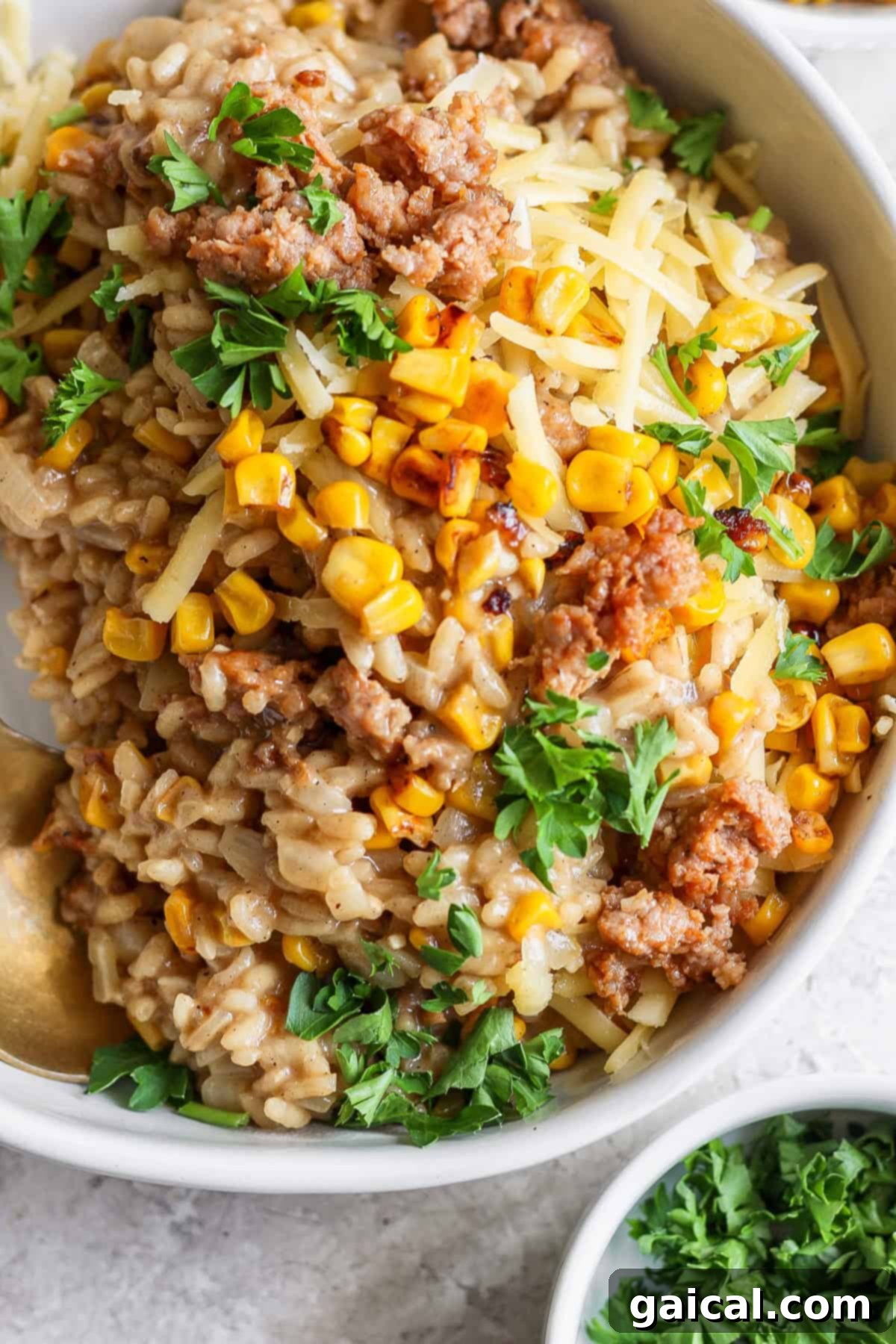 A close-up shot of a steaming bowl of creamy sweet corn risotto, generously topped with savory cooked Italian sausage, bright golden corn kernels, a sprinkle of shredded cheese, and fresh green parsley. A small bowl of neatly chopped parsley sits in the background, subtly out of focus.