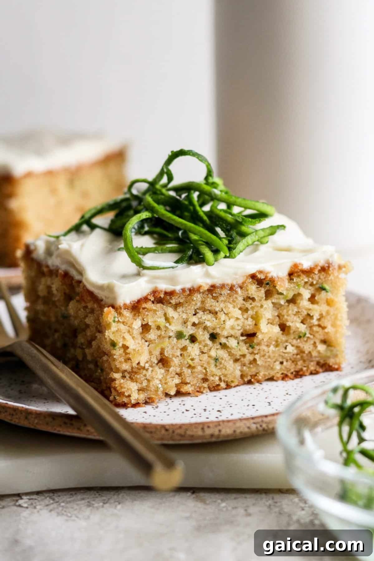 A beautifully frosted slice of zucchini cake, garnished with delicate strips of fresh zucchini, rests on a pristine white plate beside a fork, ready to be enjoyed.