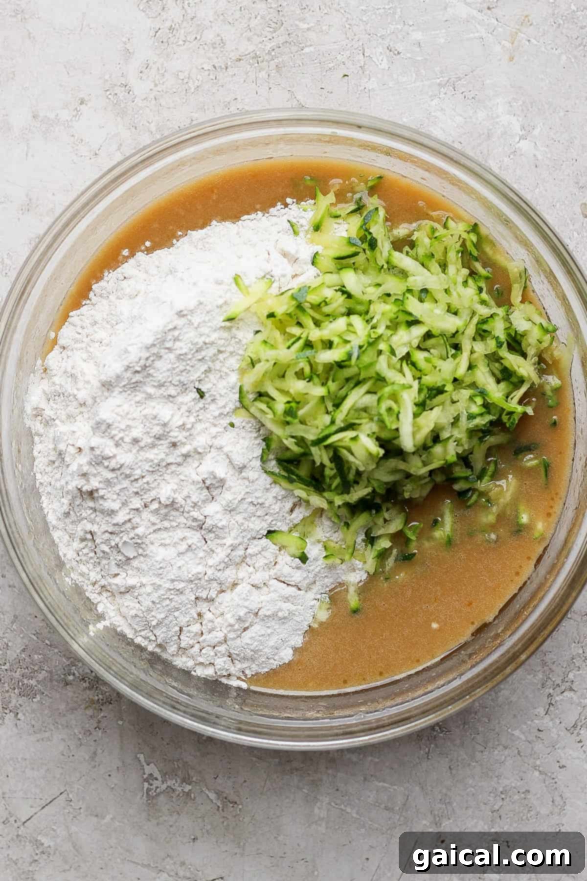 A glass mixing bowl brimming with a golden-brown cake batter, flecked with grated green zucchini, sits on a light gray countertop, surrounded by baking essentials.