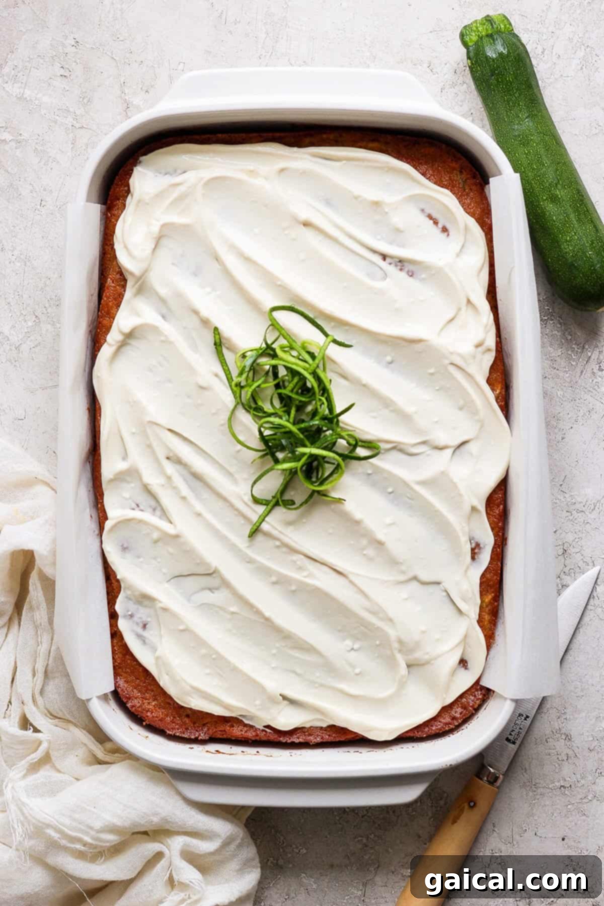 A delectable frosted zucchini sheet cake, elegantly garnished with spiralized zucchini, rests within a rectangular ceramic baking dish. Adjacent to it, a knife, a neatly folded cloth, and a whole fresh zucchini complete the inviting culinary scene.