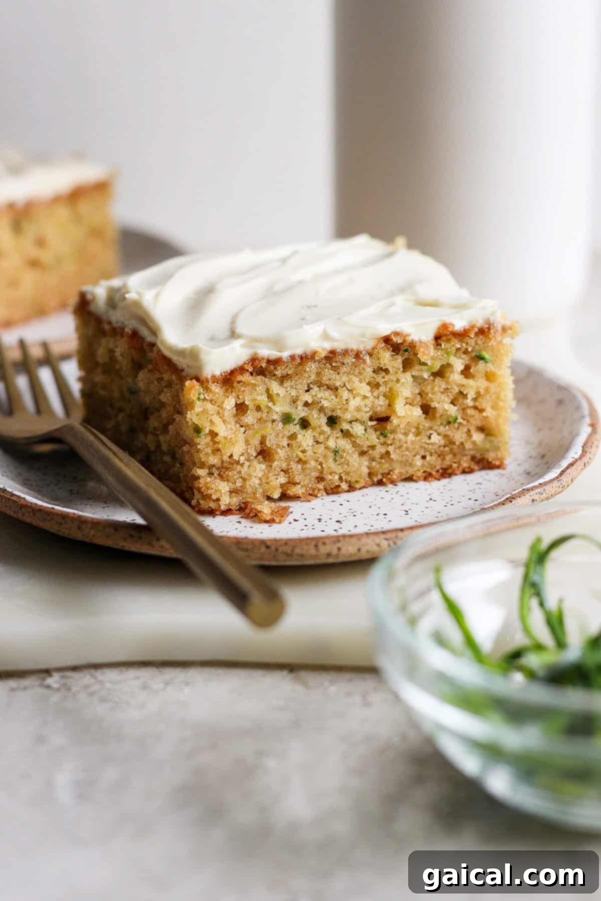 A perfectly portioned slice of frosted zucchini cake, adorned with green zest, sits invitingly on a speckled plate. A fork rests beside it, while a small bowl of fresh green garnish is softly blurred in the foreground, hinting at fresh flavors.