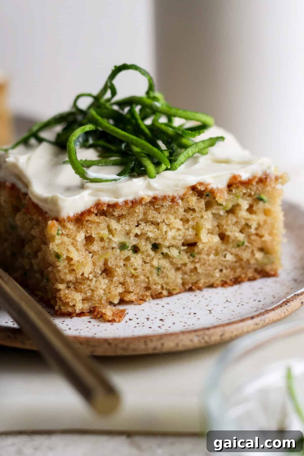 A close-up of a delightful slice of zucchini cake, crowned with smooth white frosting and garnished with vibrant green zest, presented on a chic speckled plate next to a silver fork.