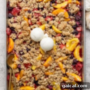 A rectangular baking tray filled with a mixed berry and peach crumble topped with two scoops of vanilla ice cream. A gold spoon rests on the left side of the tray, indicating it's ready to be served.
