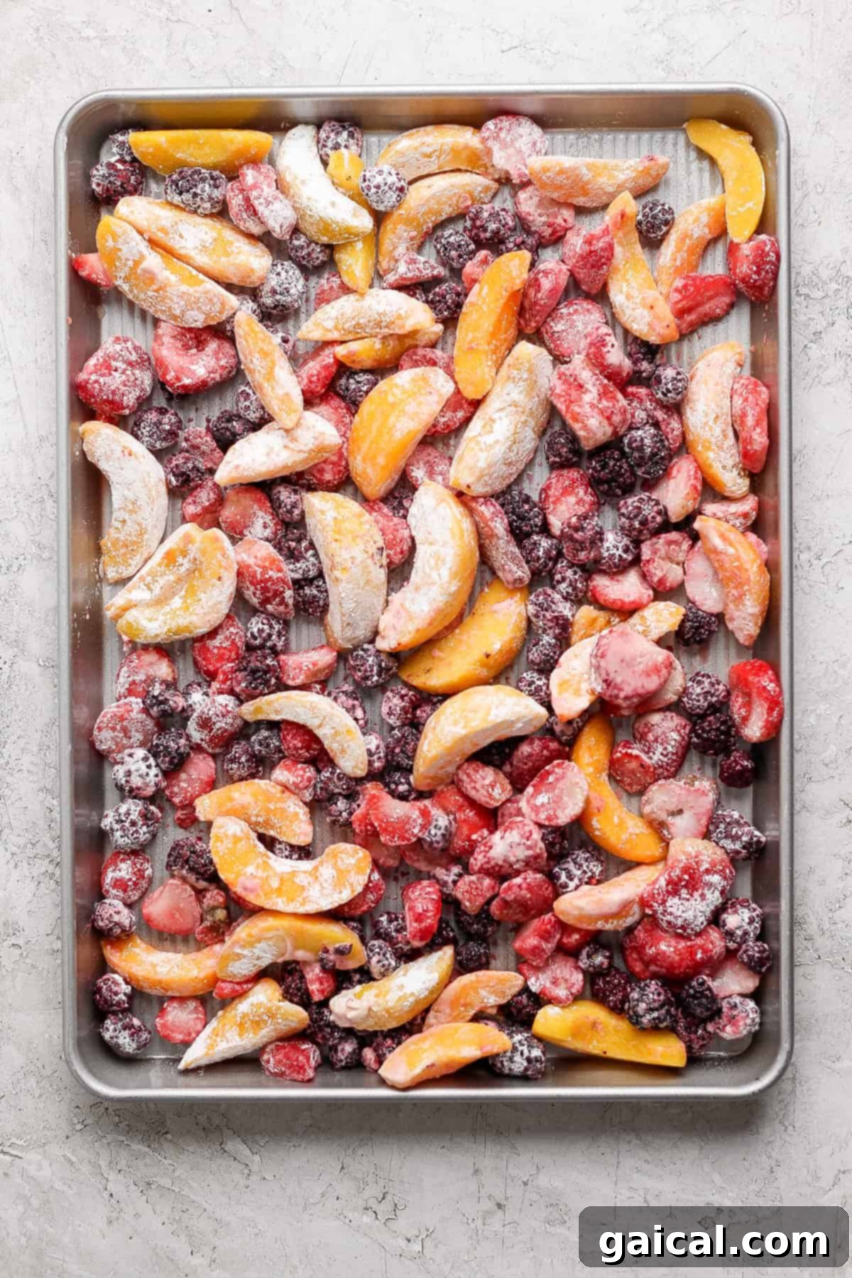 A baking sheet filled with an assortment of frozen fruit slices including peaches, strawberries, and blackberries arranged evenly across the surface, ready for the crumble topping.