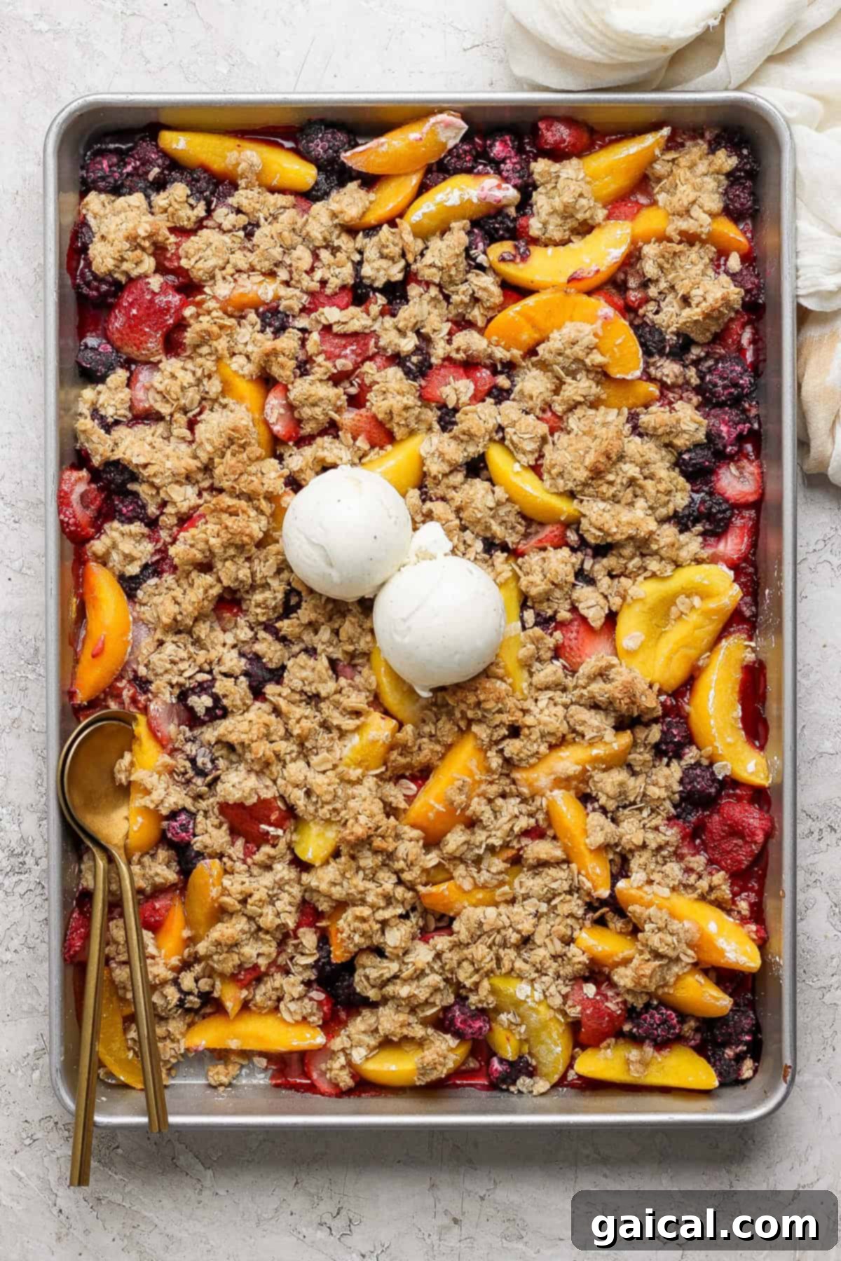 A rectangular baking tray filled with a mixed berry and peach crumble topped with two scoops of vanilla ice cream. A gold spoon rests on the left side of the tray.