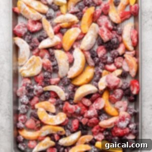 A baking sheet filled with an assortment of frozen fruit slices including peaches, strawberries, and blackberries arranged evenly across the surface, coated in cornstarch, ready for the crumble topping.