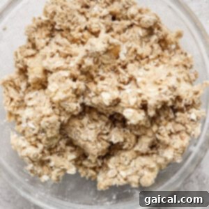 A glass bowl containing a mixture of rolled oats, flour, and brown sugar, being mixed with softened butter to form a crumbly topping.