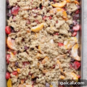 A baking tray filled with a vibrant mixture of sliced peaches, strawberries, and blackberries, generously topped with a golden, crumbly oat mixture, ready to be baked to perfection.