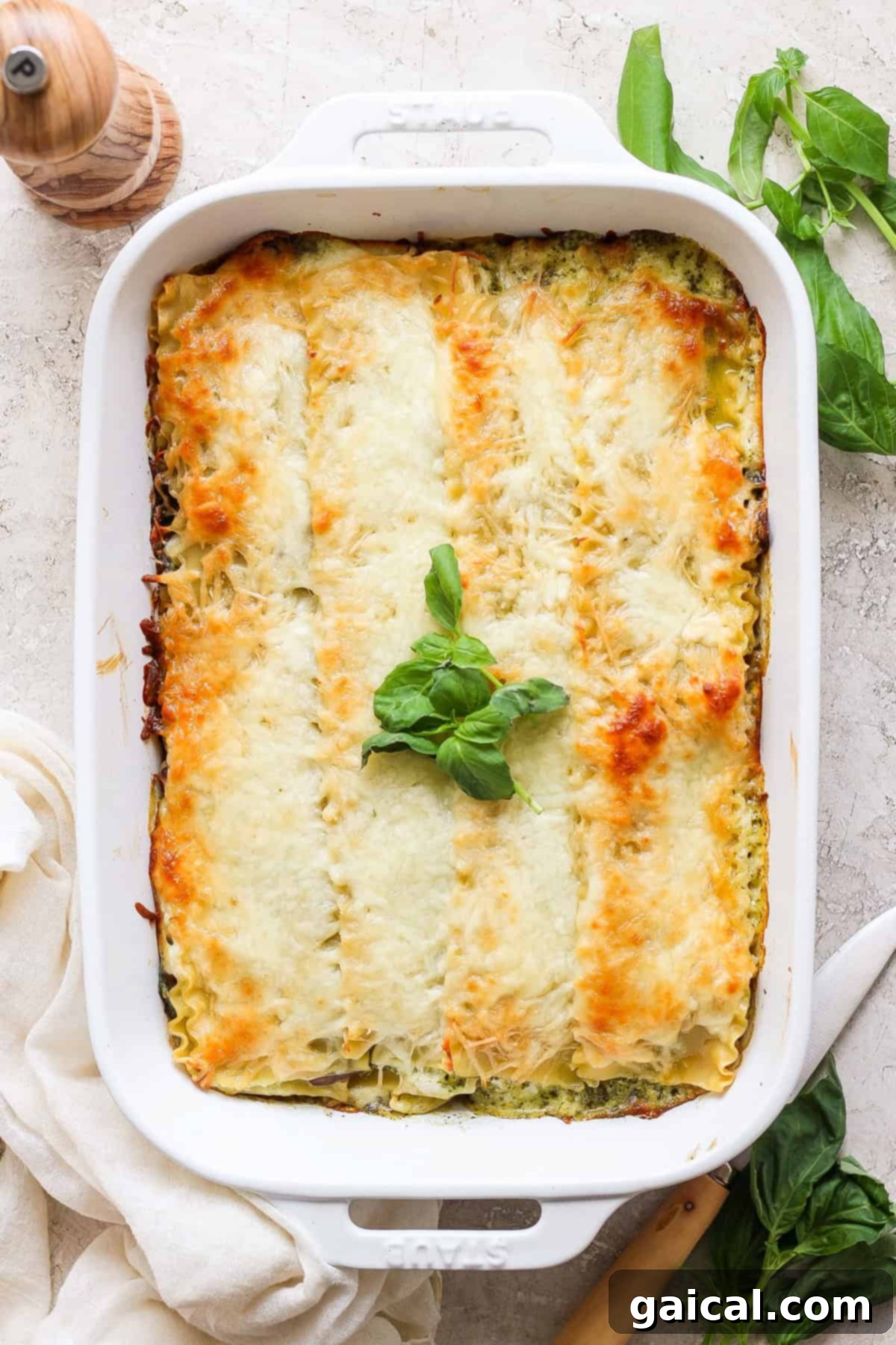 Rustic Pesto Lasagna 5 A beautifully baked pesto lasagna in a white rectangular dish, topped with golden-brown melted cheese and artfully garnished with fresh basil leaves. In the background, a wooden pepper grinder and more fresh basil suggest a delightful dining experience.