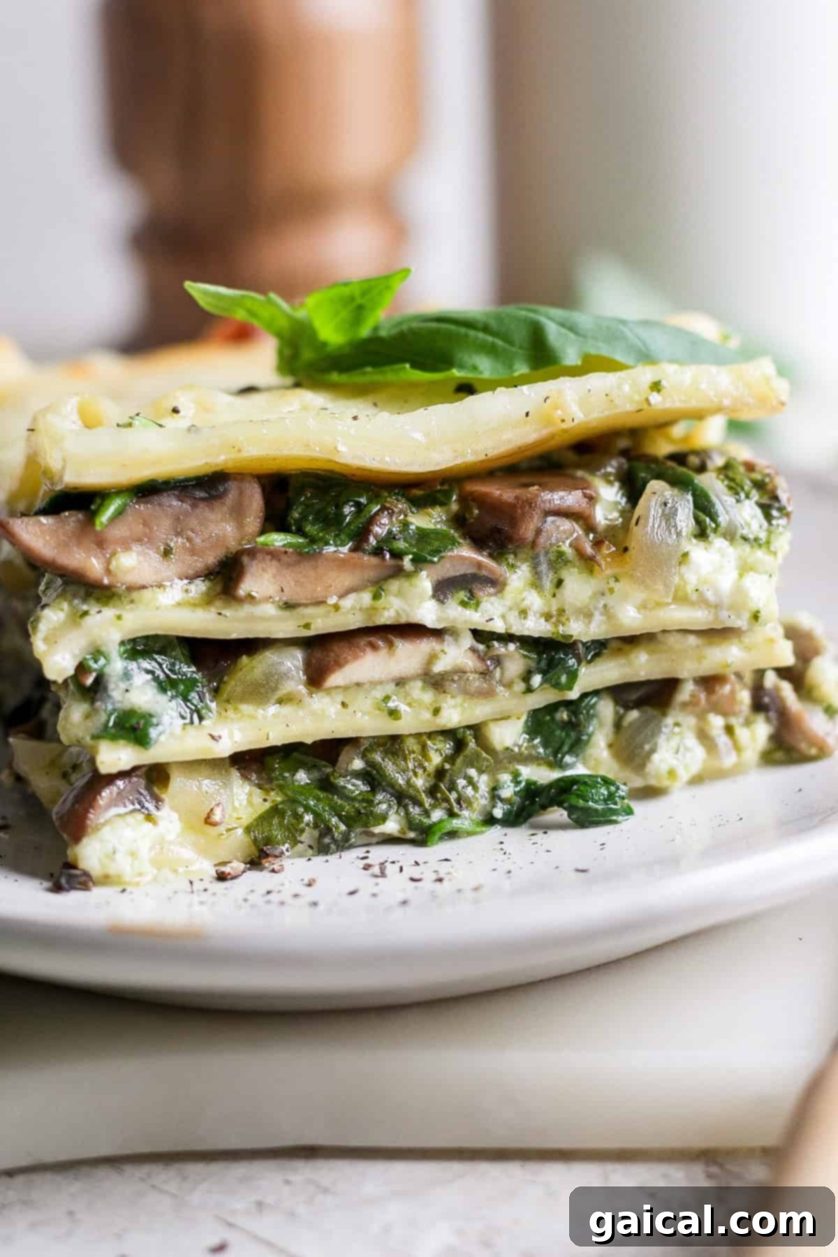 Rustic Pesto Lasagna 8 A delectable slice of vegetarian lasagna resting on a crisp white plate, showcasing its vibrant layers of pasta, creamy cheese, rich mushrooms, and fresh spinach, elegantly topped with a single basil leaf for garnish.