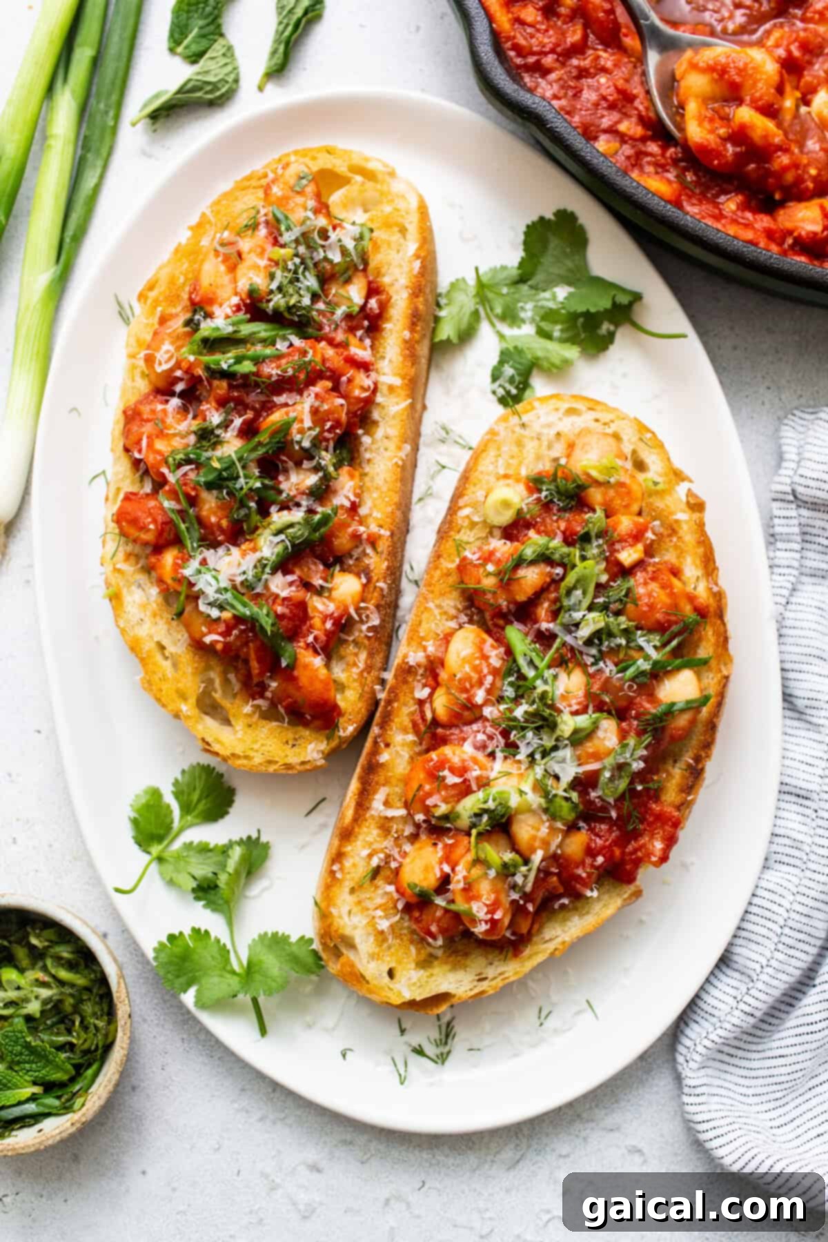 Two slices of hearty toast generously topped with a rich butter bean and tomato mixture, garnished with fresh green herbs and grated Parmesan cheese, presented on a rustic plate.