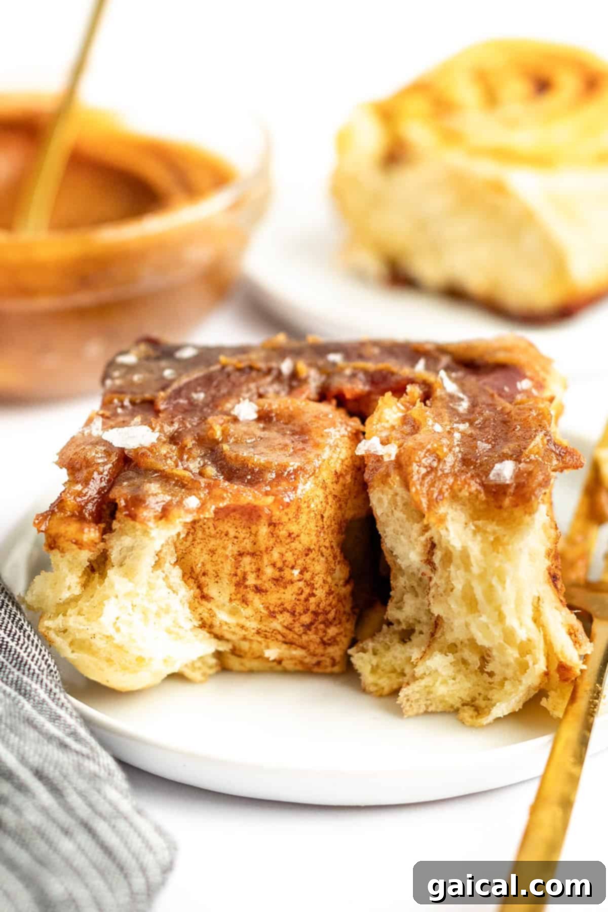 Gooey Cinnamon Apple Rolls 2 Close-up of a cinnamon roll with glaze and a sprinkle of salt on a white plate. A partially eaten cinnamon roll and a dish with more glaze are in the background.