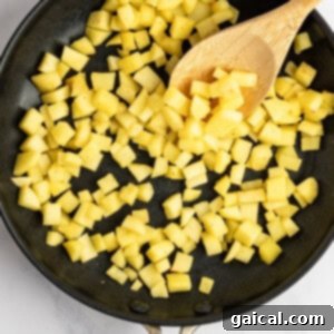 Gooey Cinnamon Apple Rolls 11 Diced potatoes cooking in a black skillet with a wooden spoon on a white marble surface.