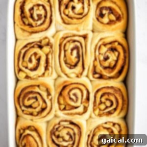 Gooey Cinnamon Apple Rolls 16 A rectangular baking dish filled with twelve freshly baked cinnamon rolls, each with a visible swirl pattern, on a white surface.
