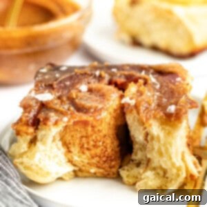 Gooey Cinnamon Apple Rolls 18 Close-up of a cinnamon roll with glaze and a sprinkle of salt on a white plate. A partially eaten cinnamon roll and a dish with more glaze are in the background.