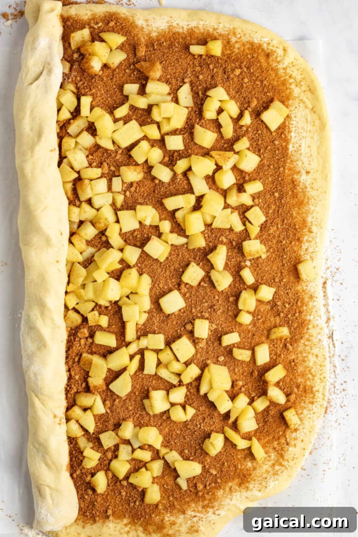 Gooey Cinnamon Apple Rolls 4 Unbaked cinnamon roll dough topped with diced apples and cinnamon sugar, partially rolled up on a light surface.