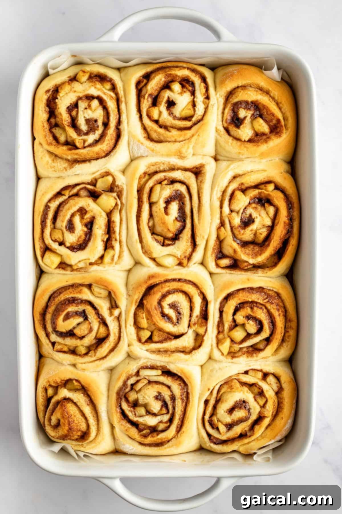 Gooey Cinnamon Apple Rolls 5 A rectangular baking dish filled with twelve freshly baked cinnamon rolls, each with a visible swirl pattern, on a white surface.