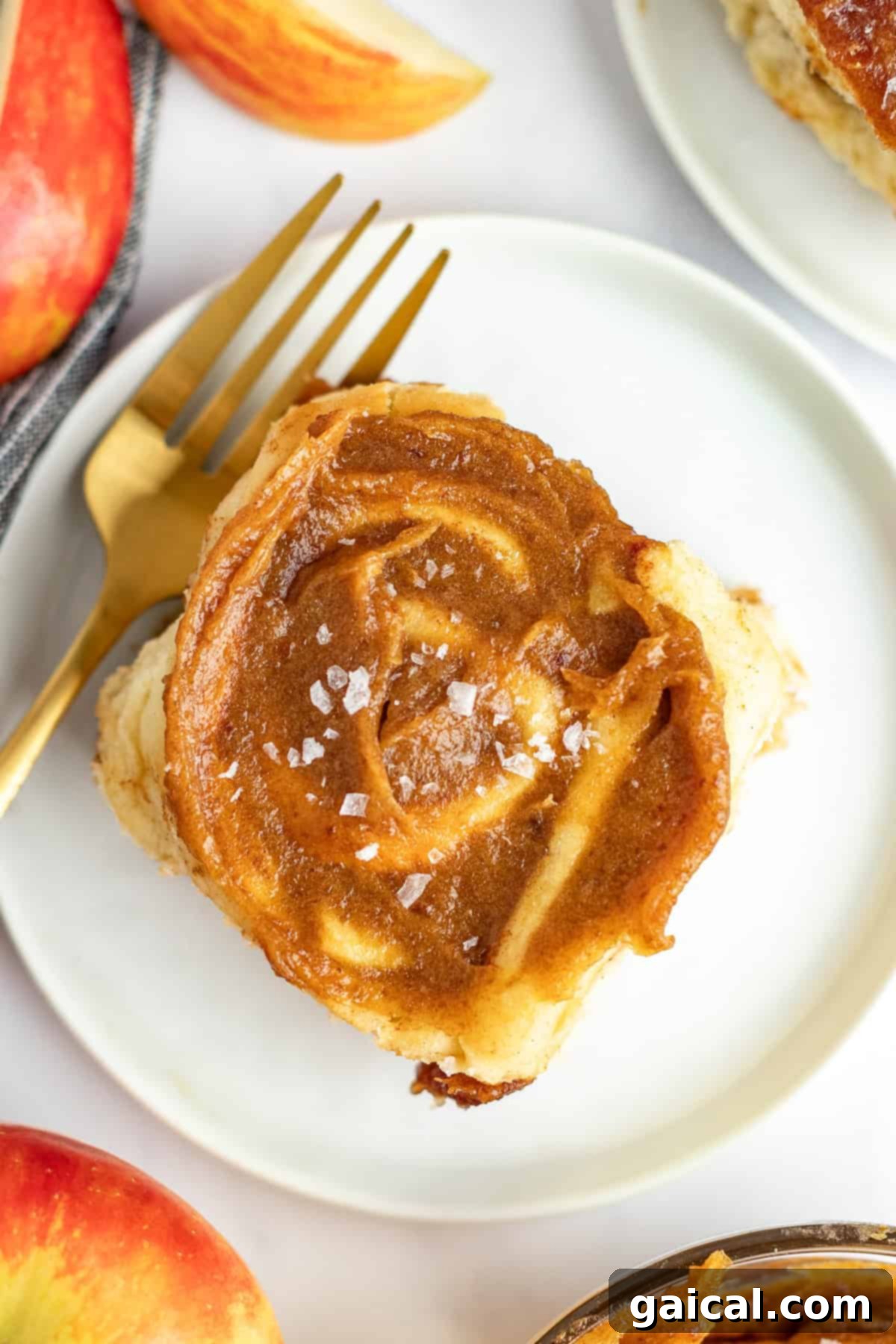 Gooey Cinnamon Apple Rolls 7 A single cinnamon roll with frosting and a sprinkle of sea salt is on a white plate with a gold fork. Slices of a red apple are partially visible in the background.