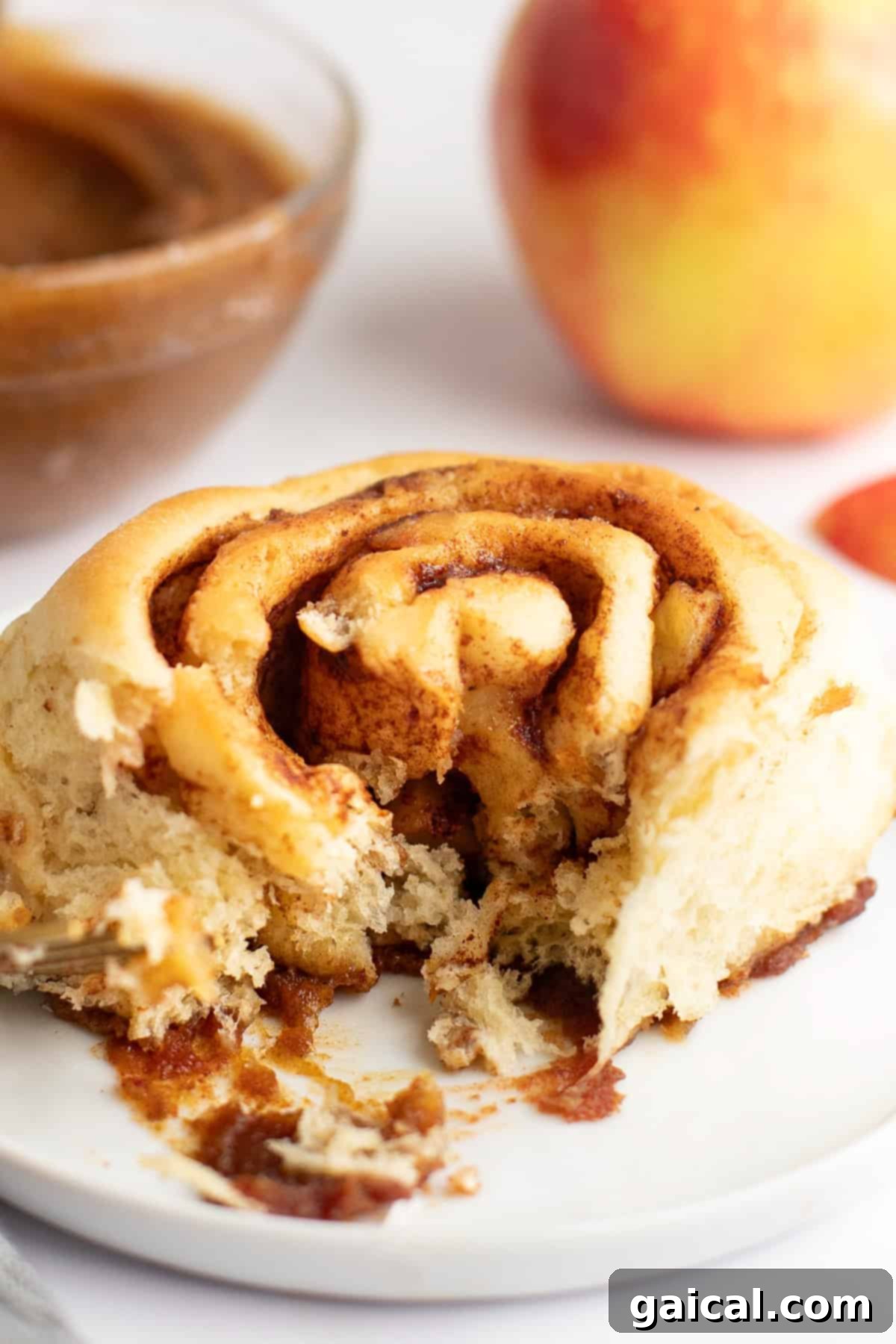Gooey Cinnamon Apple Rolls 8 A partially eaten cinnamon roll on a white plate with a bowl of filling and an apple in the background.