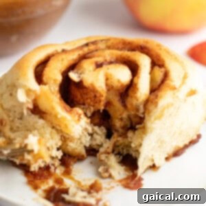 Gooey Cinnamon Apple Rolls 9 A close-up view of a partially eaten cinnamon roll on a white plate, showing its swirled layers and a bite taken out, with blurred background elements.