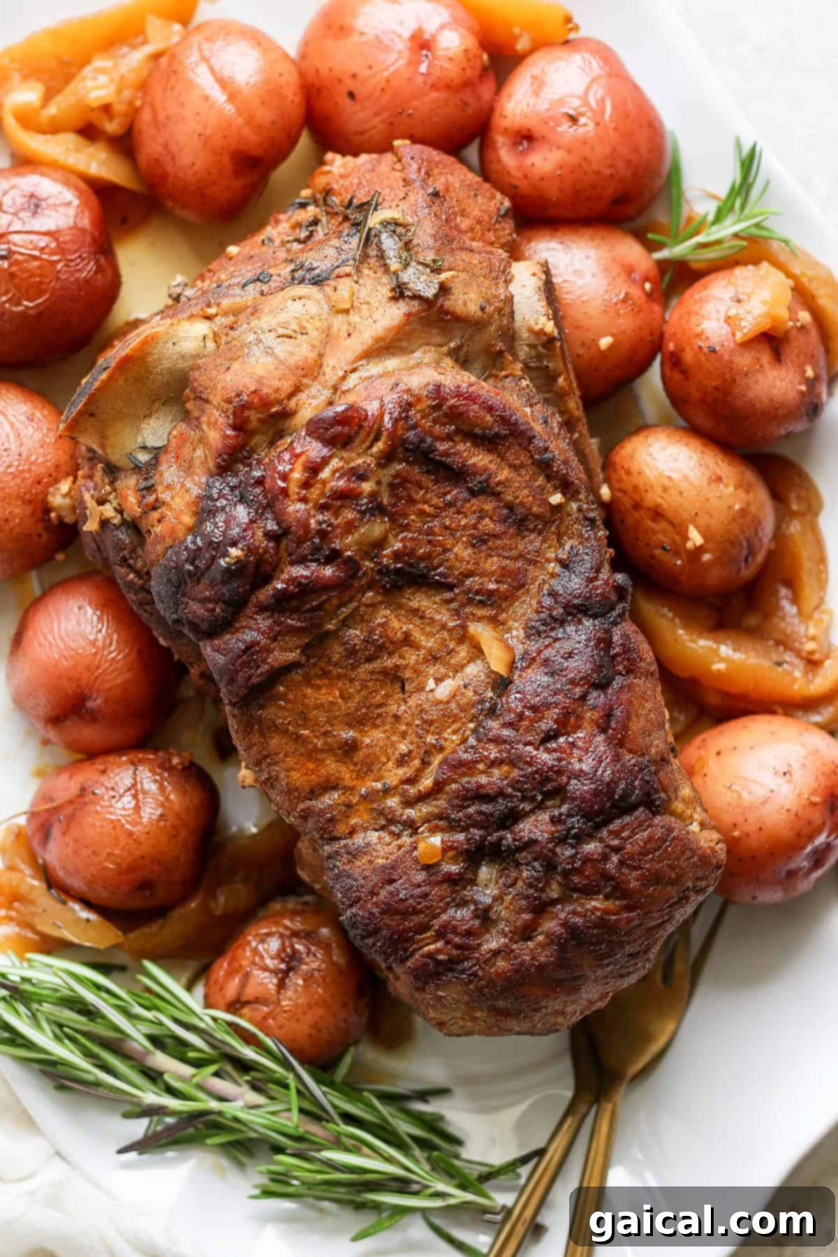 A roasted pork shoulder with a crispy exterior is surrounded by browned red potatoes and sprigs of rosemary on a white serving plate.