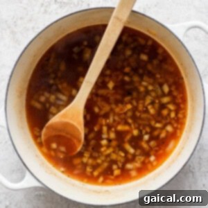 A white pot filled with a red broth containing chopped onions, stirred with a wooden spoon, placed on a gray textured surface.