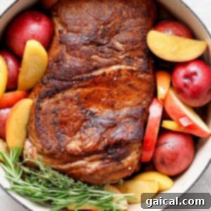 A cooked pork roast with a crispy exterior, surrounded by red potatoes, apple slices, and sprigs of rosemary in a white cast iron pot.