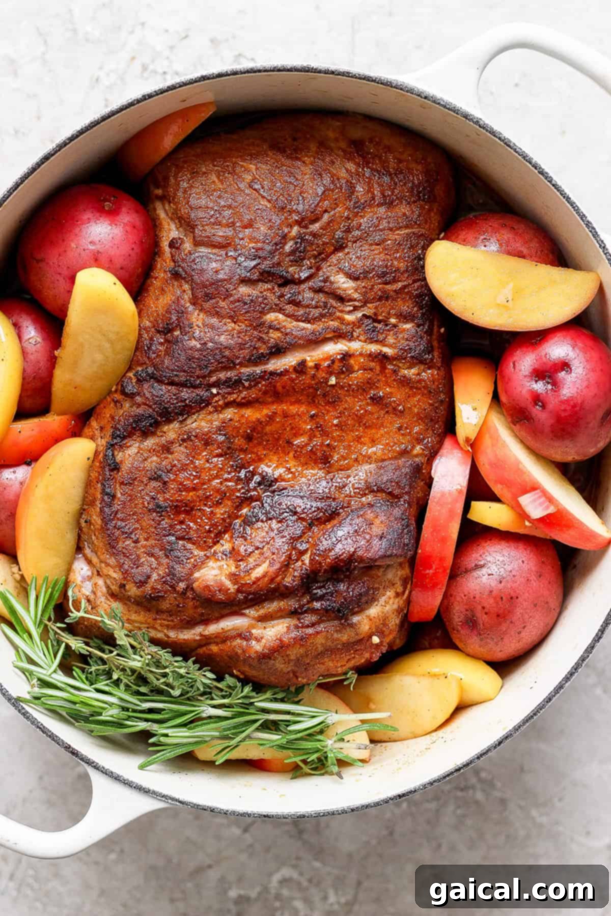 A cooked pork roast with a crispy exterior, surrounded by red potatoes, apple slices, and sprigs of rosemary in a white cast iron pot.