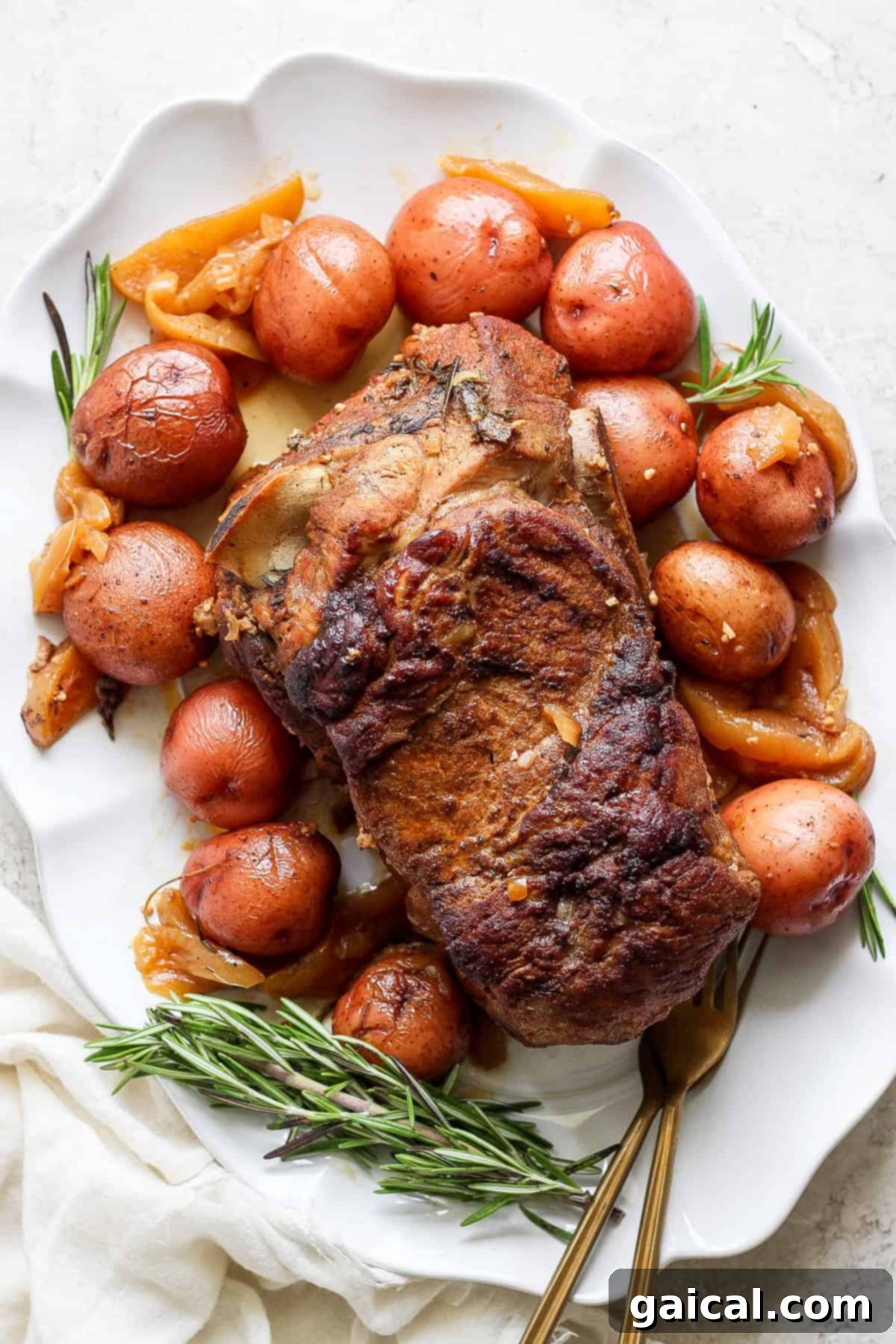 A roasted pork shoulder is served on a white plate, accompanied by red potatoes, onions, and garnished with rosemary sprigs. A white cloth is partially visible on the side.