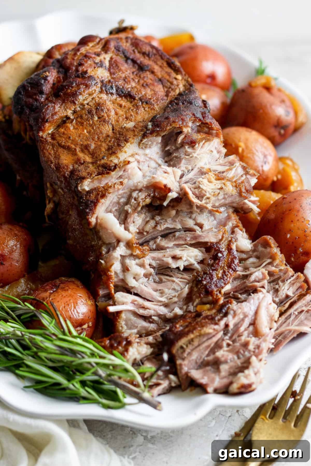 A close-up of a seasoned, cooked roast surrounded by whole red potatoes and fresh rosemary sprigs on a white platter. A fork and knife are set to the side.