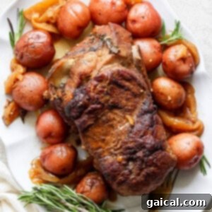 An oven-roasted pork roast surrounded by red potatoes and garnished with rosemary on a white platter.