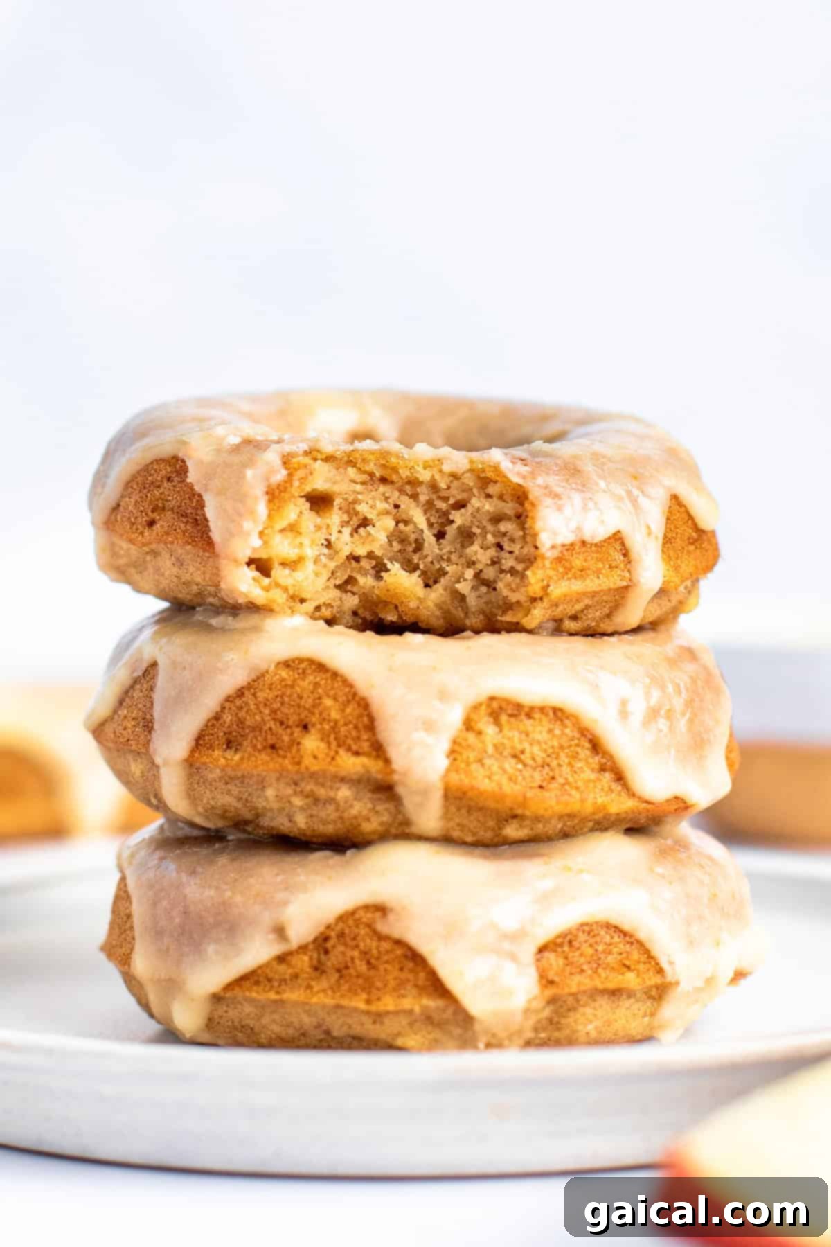 Spiced Orchard Doughnuts 2 A stack of three glazed apple cider donuts on a white plate, with the top donut partially eaten, revealing its golden brown, tender interior.