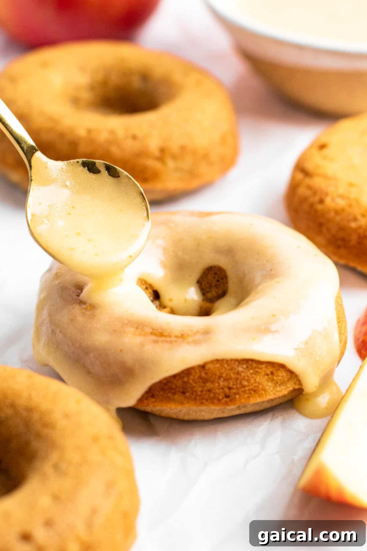 Spiced Orchard Doughnuts 6 A spoon drizzles a thick, glossy brown butter glaze over a baked apple cider donut, with more glazed donuts and a sliced apple in the blurred background.