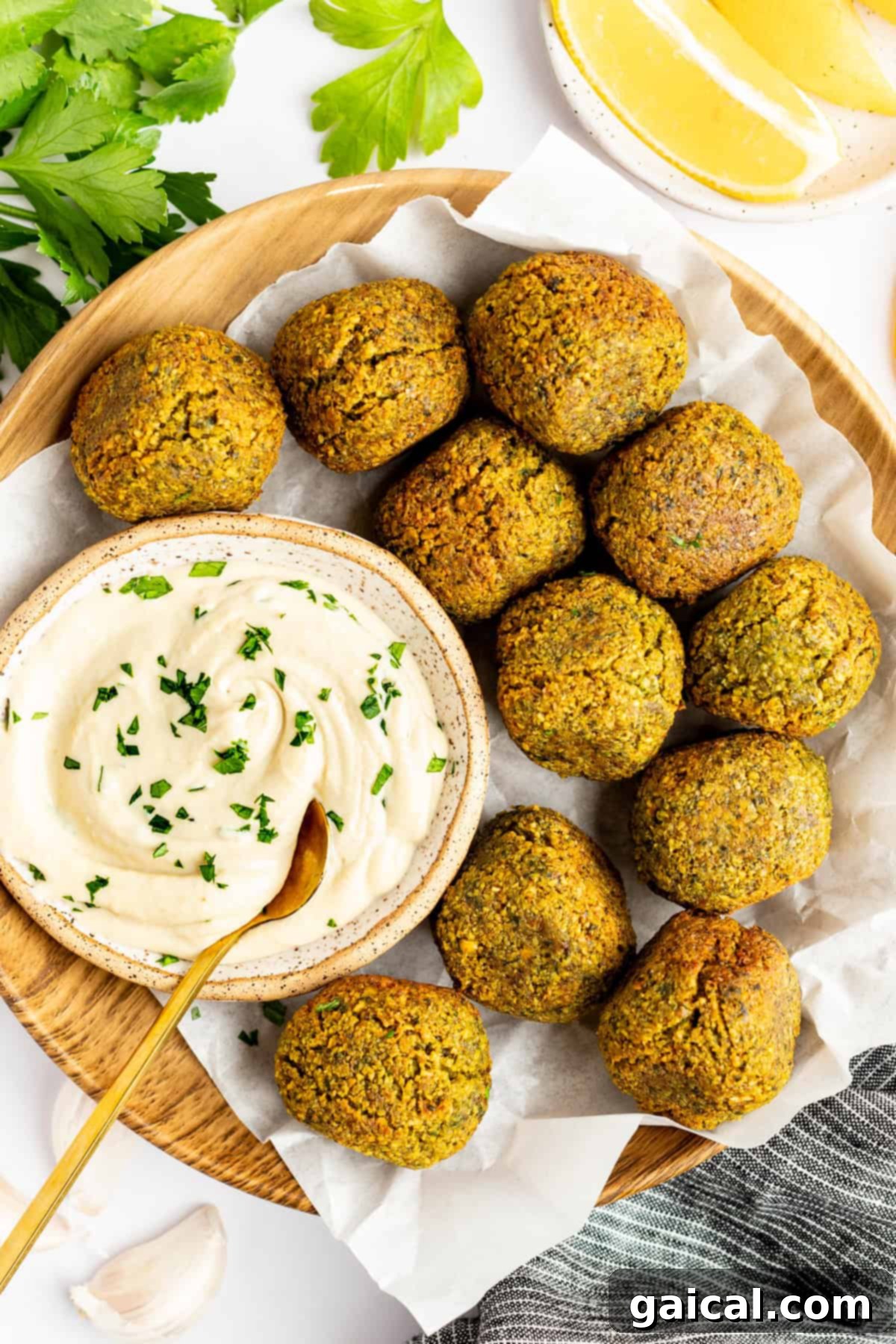 A wooden plate holds ten falafel balls surrounding a bowl of creamy dip garnished with herbs. Lemon wedges and parsley leaves are in the background.